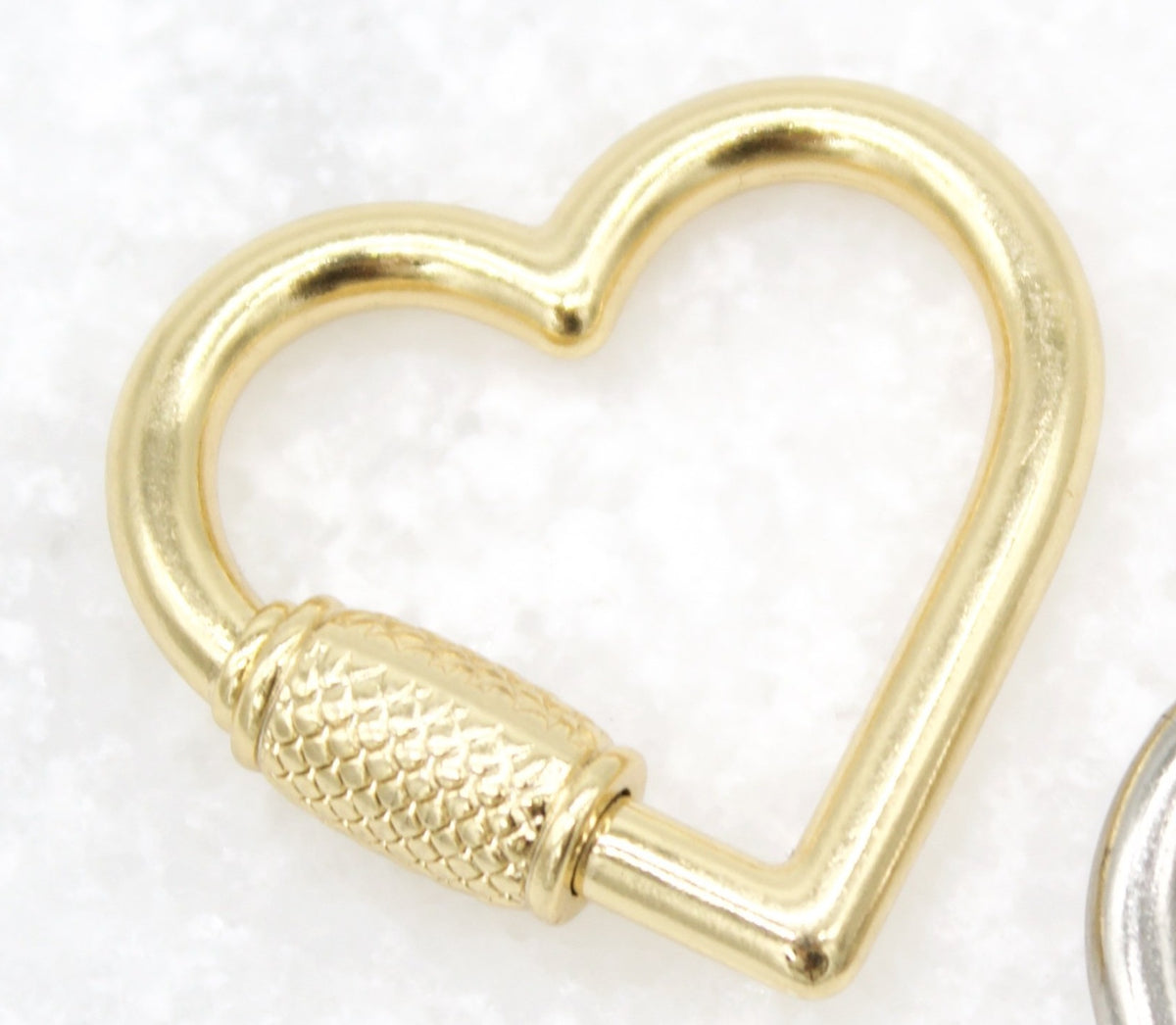 A polished gold heart-shaped carabiner features a textured screw gate closure resting on a white background.