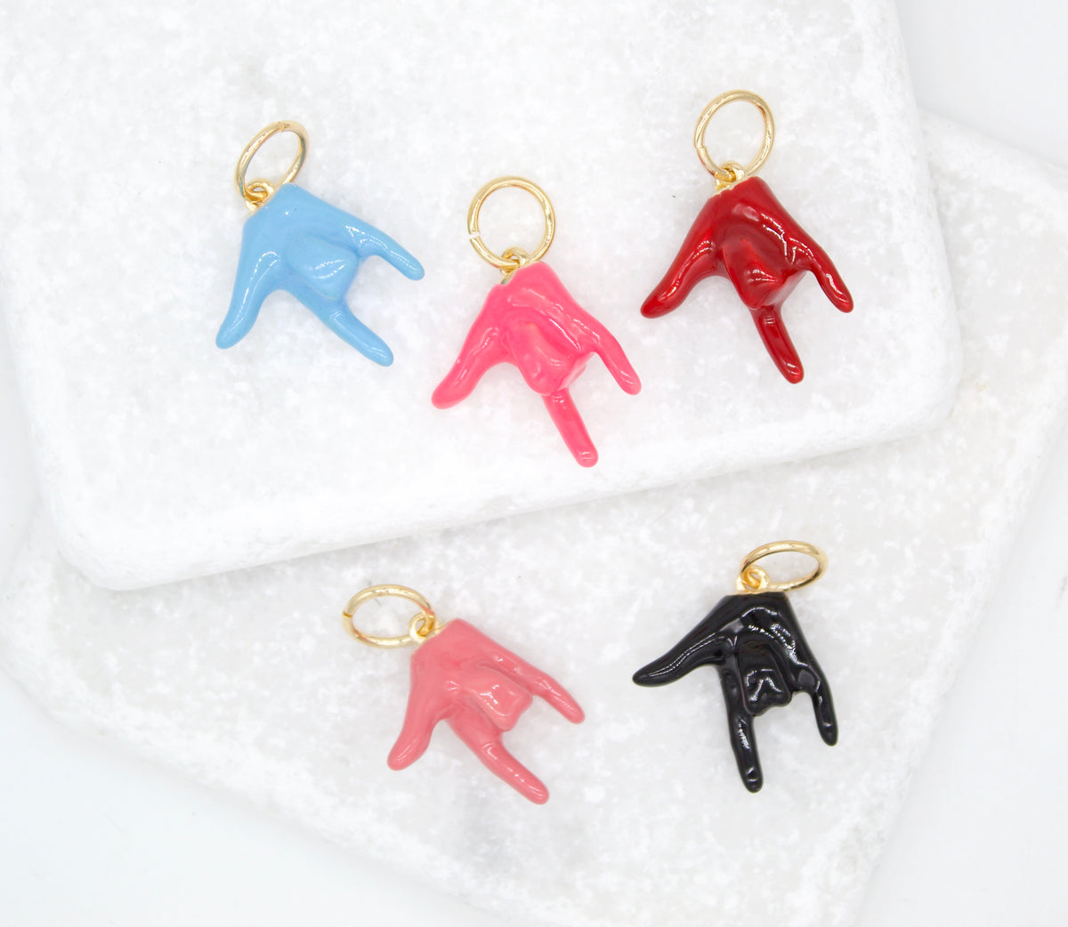 Five colorful enamel hand gesture charms in blue, pink, red, and black rest on a white marble surface.