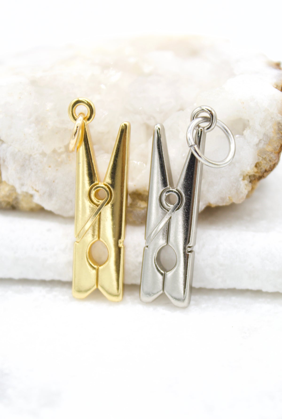 Two metallic clothespin charms, one gold and one silver, rest upright on a white crystal geode surface.