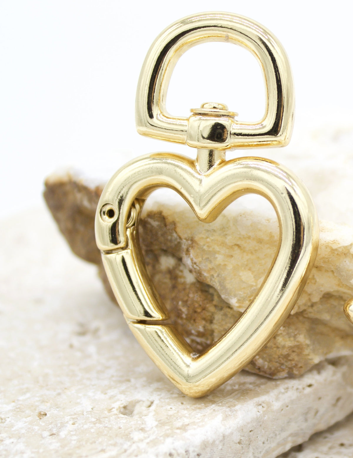 A shiny gold heart-shaped swivel lobster clasp rests on a textured stone surface.