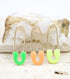 Three gold safety pin charms with colorful enamel details in green, peach, and yellow rest against a textured white crystal surface.