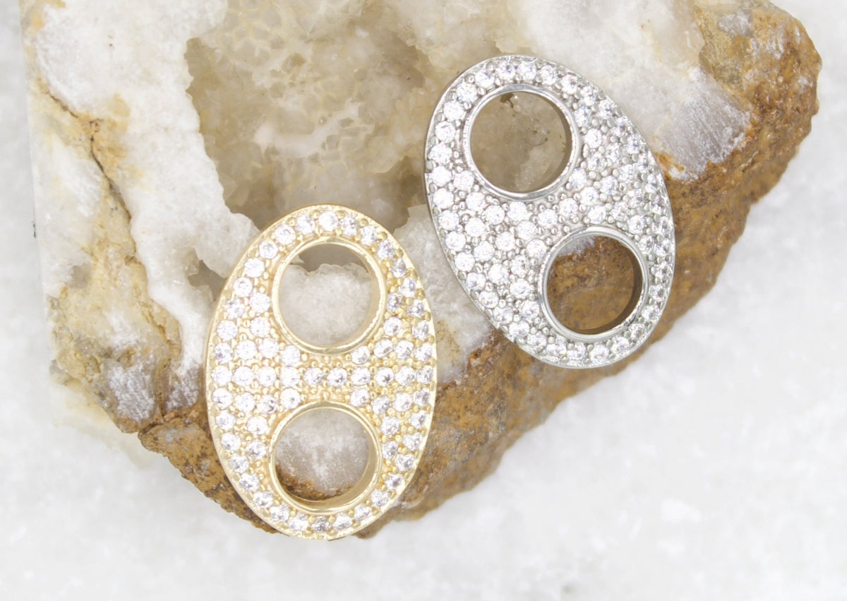 Two oval gold and silver pendant bails encrusted with sparkling cubic zirconia rest against a textured geode background.