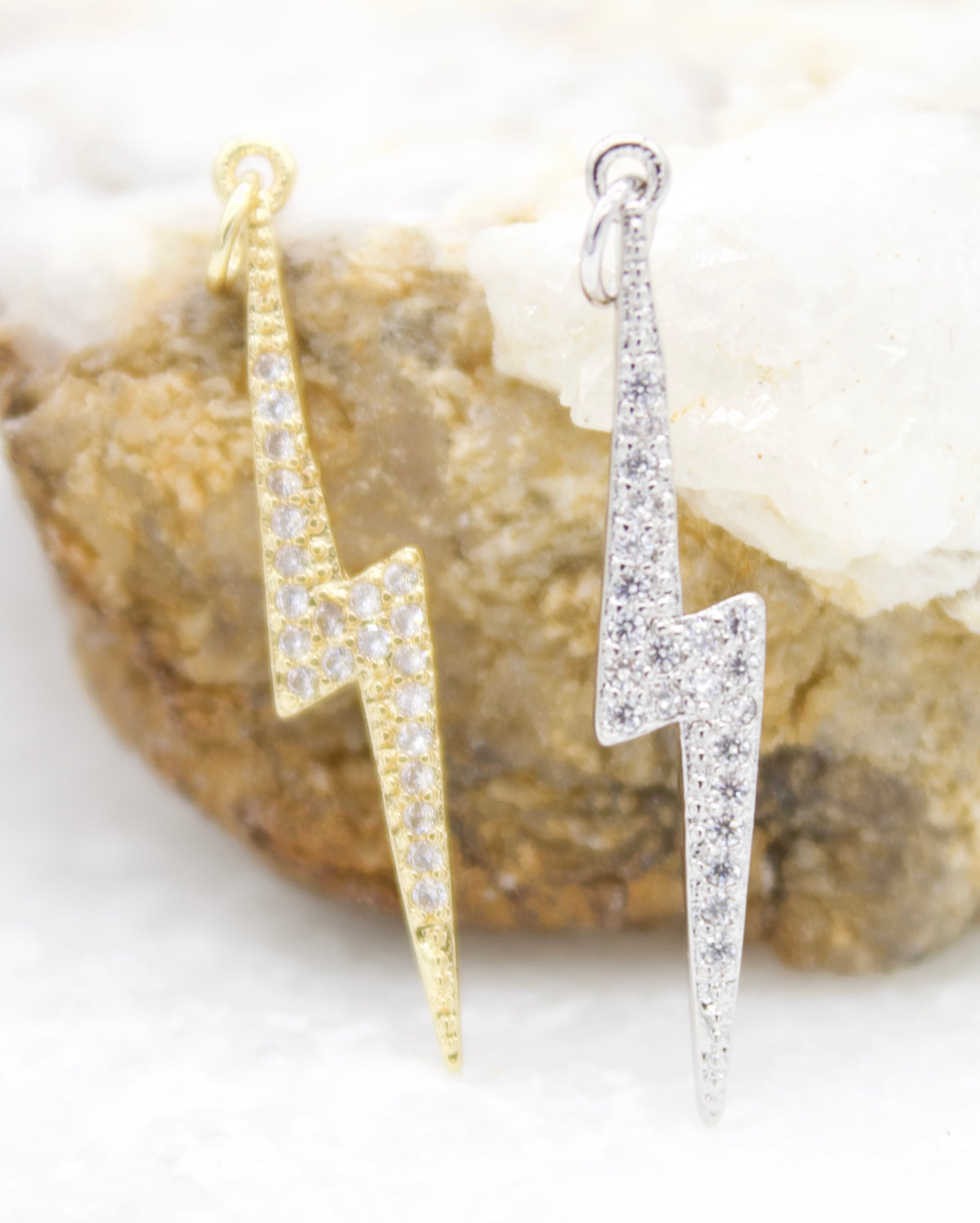 Two pavé diamond lightning bolt pendant charms, one gold and one silver, rest against a textured stone background.