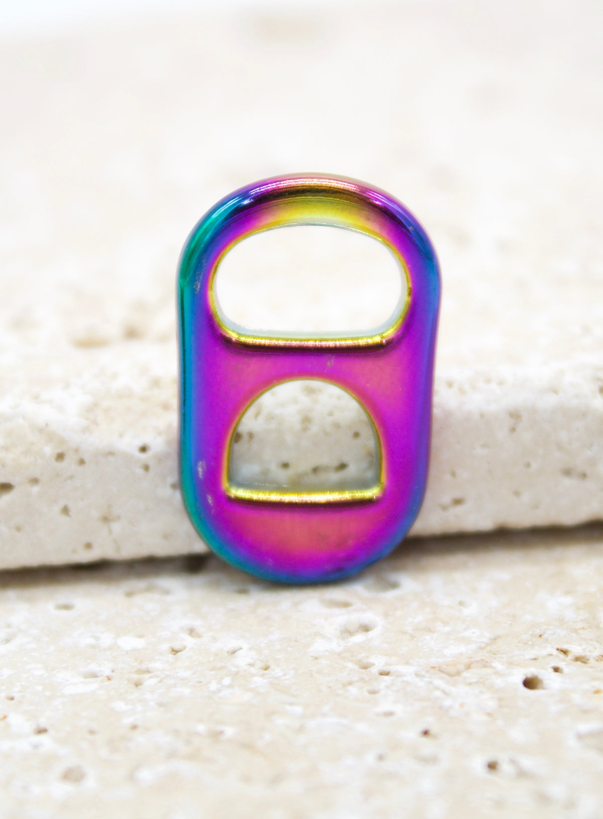 A vibrant iridescent rainbow soda tab rests upright on a textured stone surface.