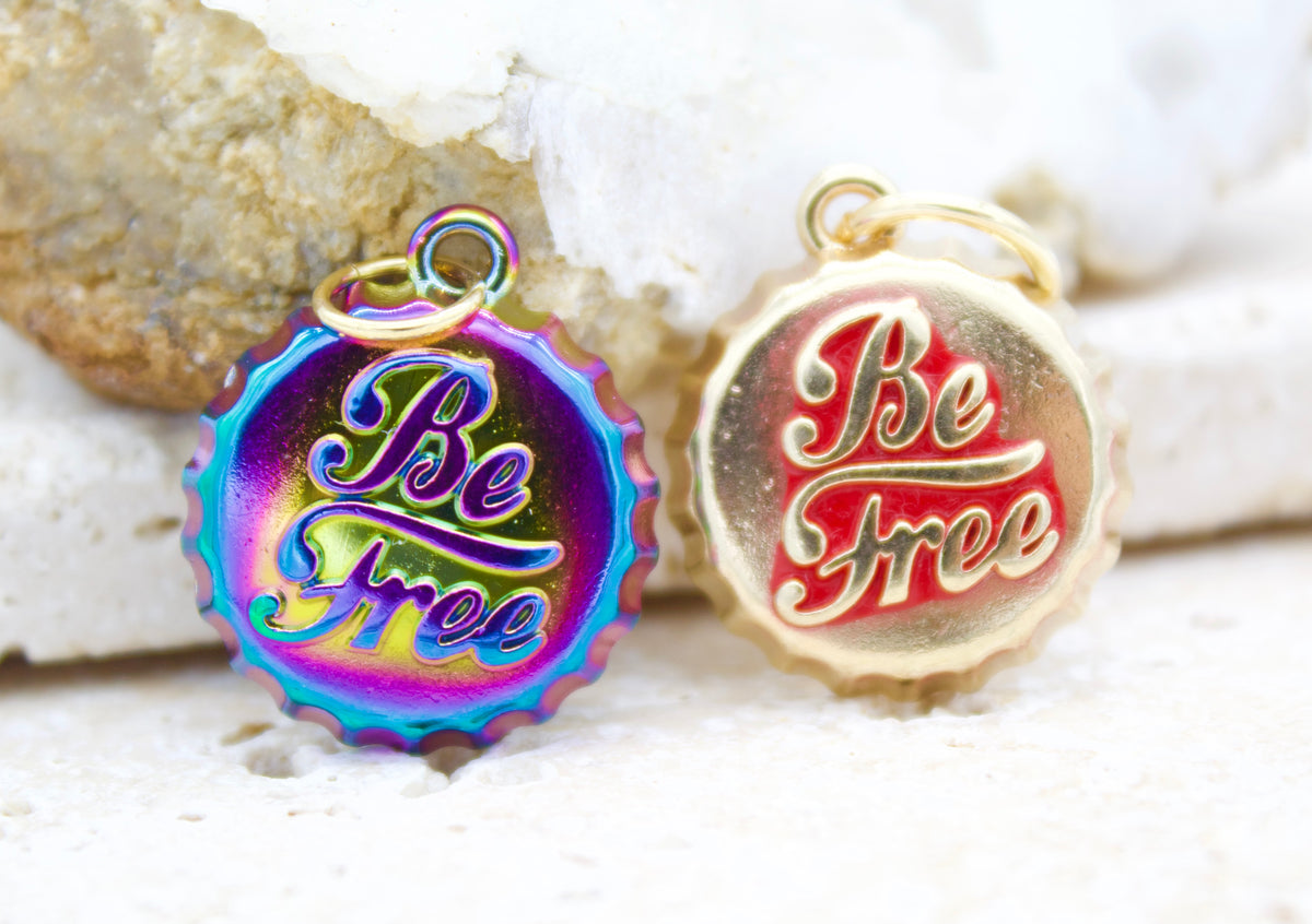 Two round "Be Free" bottle cap charms rest against a textured rock background, featuring one in iridescent rainbow and the other in gold with a red center.