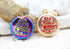 Two round "Be Free" bottle cap charms rest against a textured rock background, featuring one in iridescent rainbow and the other in gold with a red center.