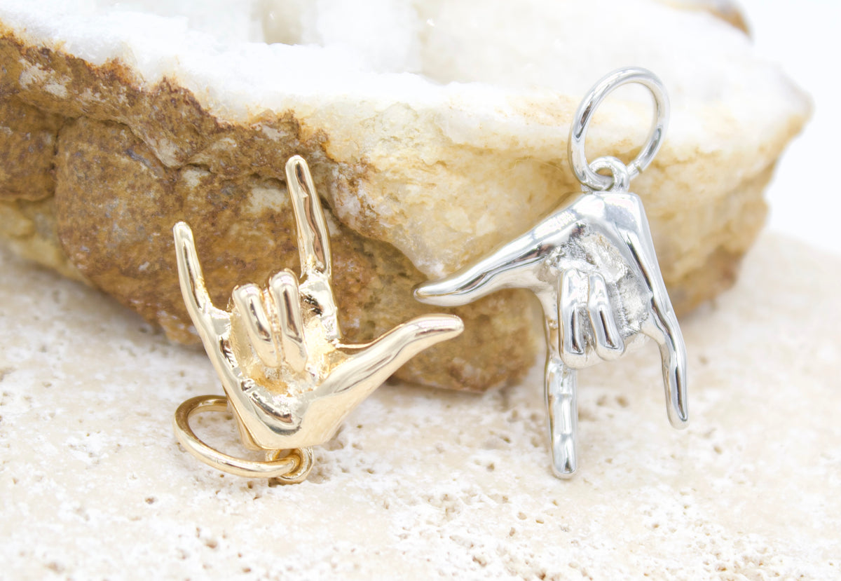 A gold and a silver American Sign Language "I love you" hand sign charm rest side-by-side against a textured crystal background.