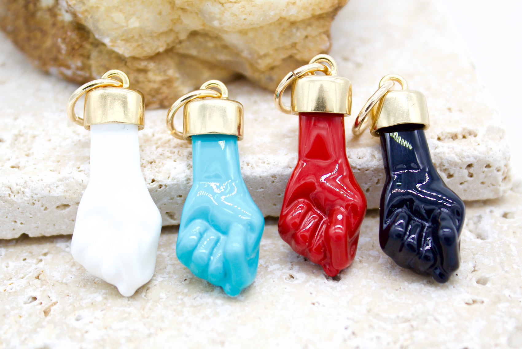 Four colorful fist pendants in white, turquoise, red, and black rest on a textured stone surface.