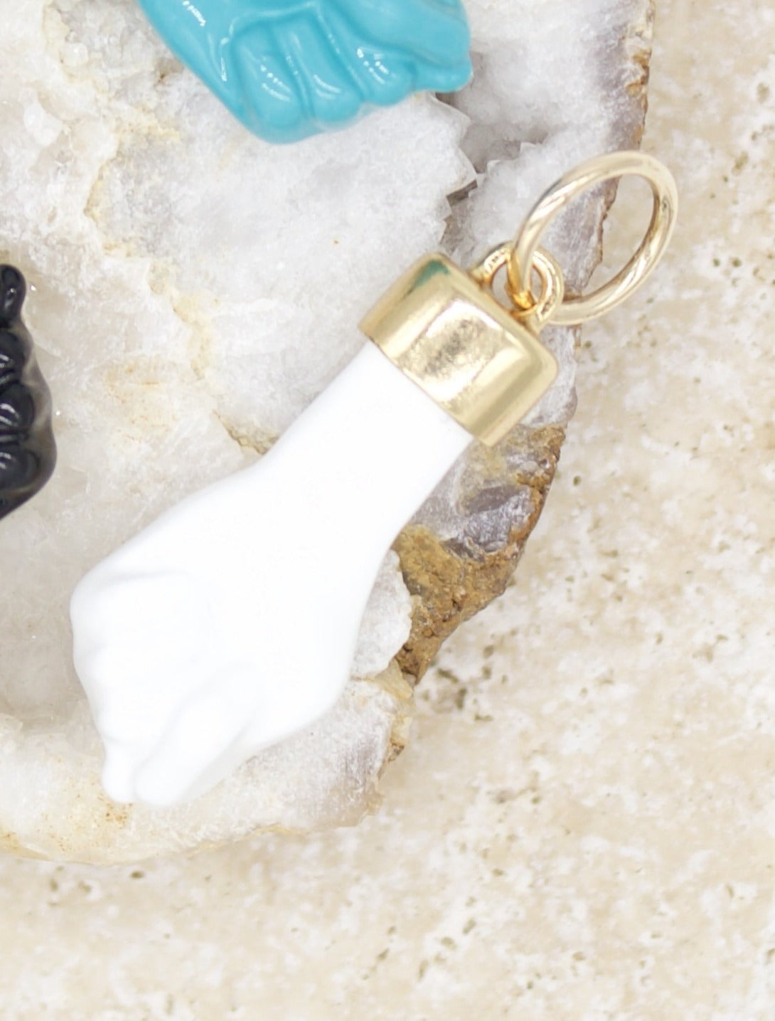 A white fist charm with a gold bail rests on a textured geode surface next to a turquoise charm.