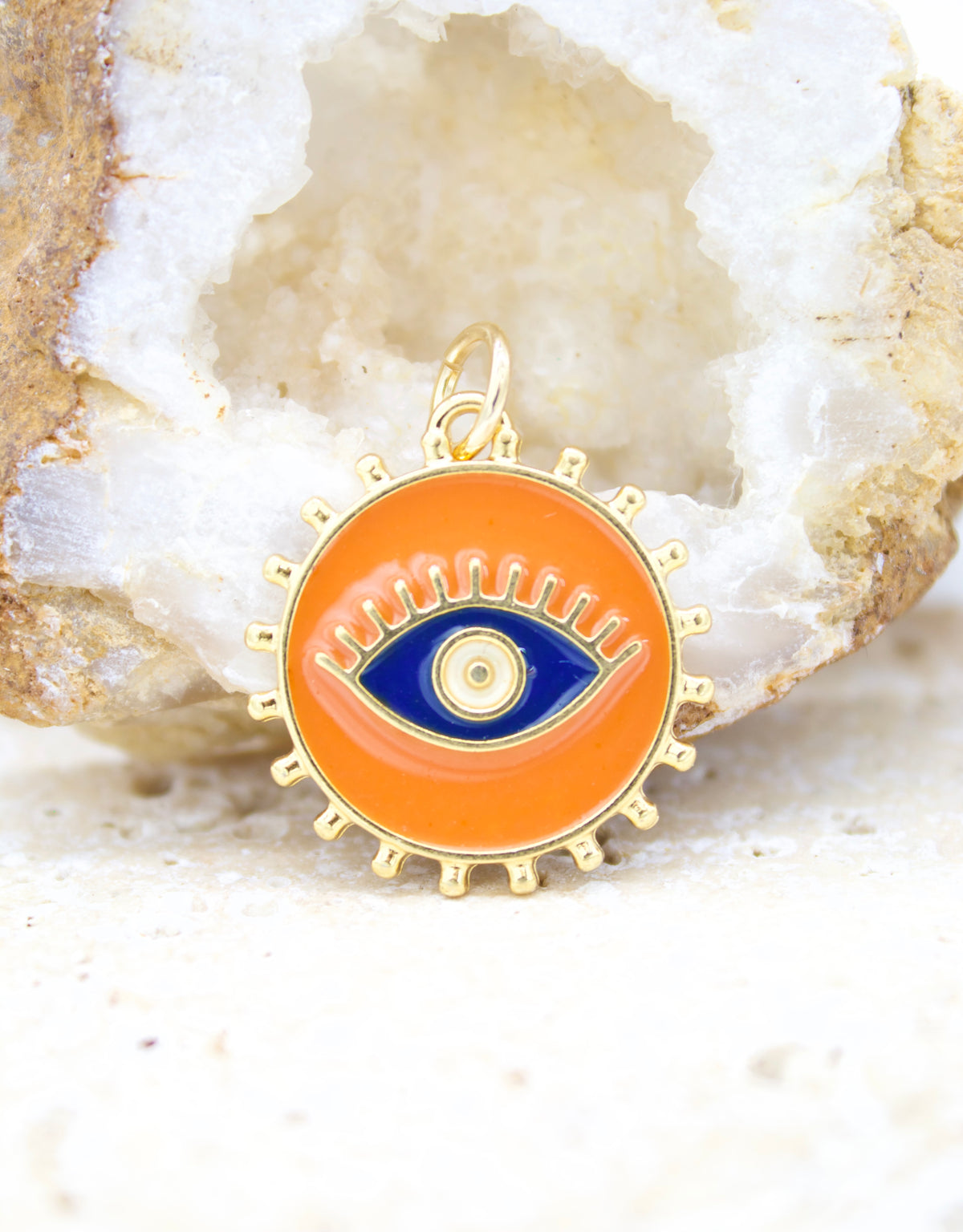 A round gold evil eye pendant featuring vibrant orange enamel and a blue eye center rests against a sparkling crystal geode.