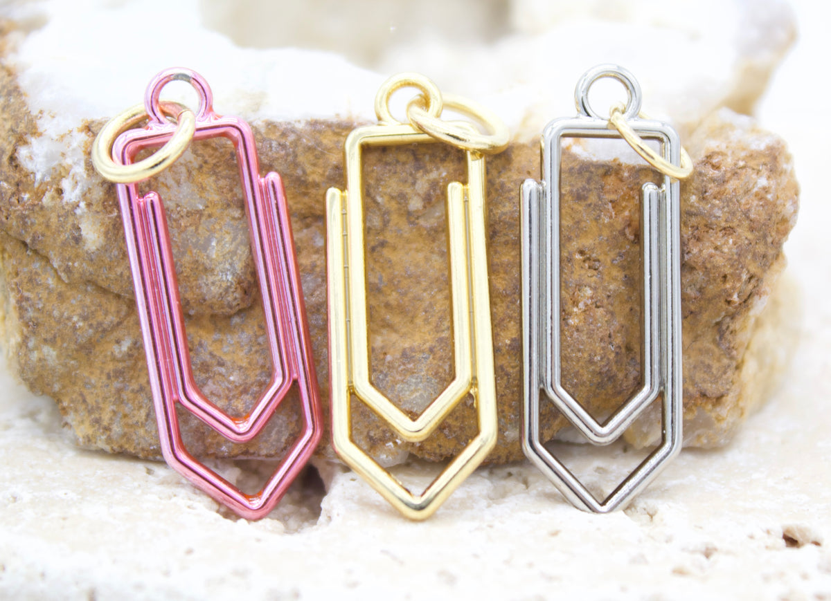 Three colorful paperclip charms in pink, gold, and silver display jump rings and rest upright against a textured stone backdrop.