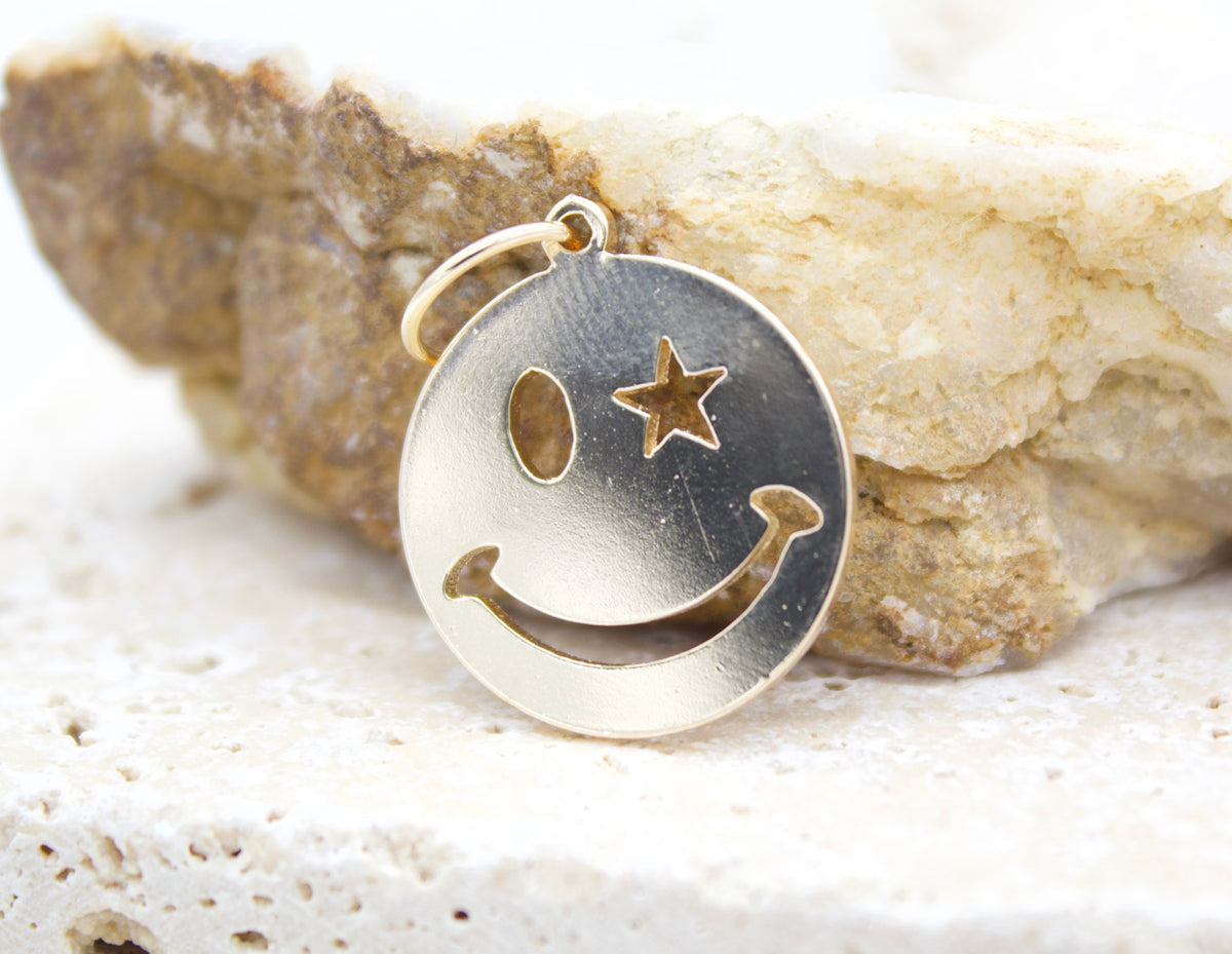 A gold smiley face pendant with a star eye rests on a textured stone surface.