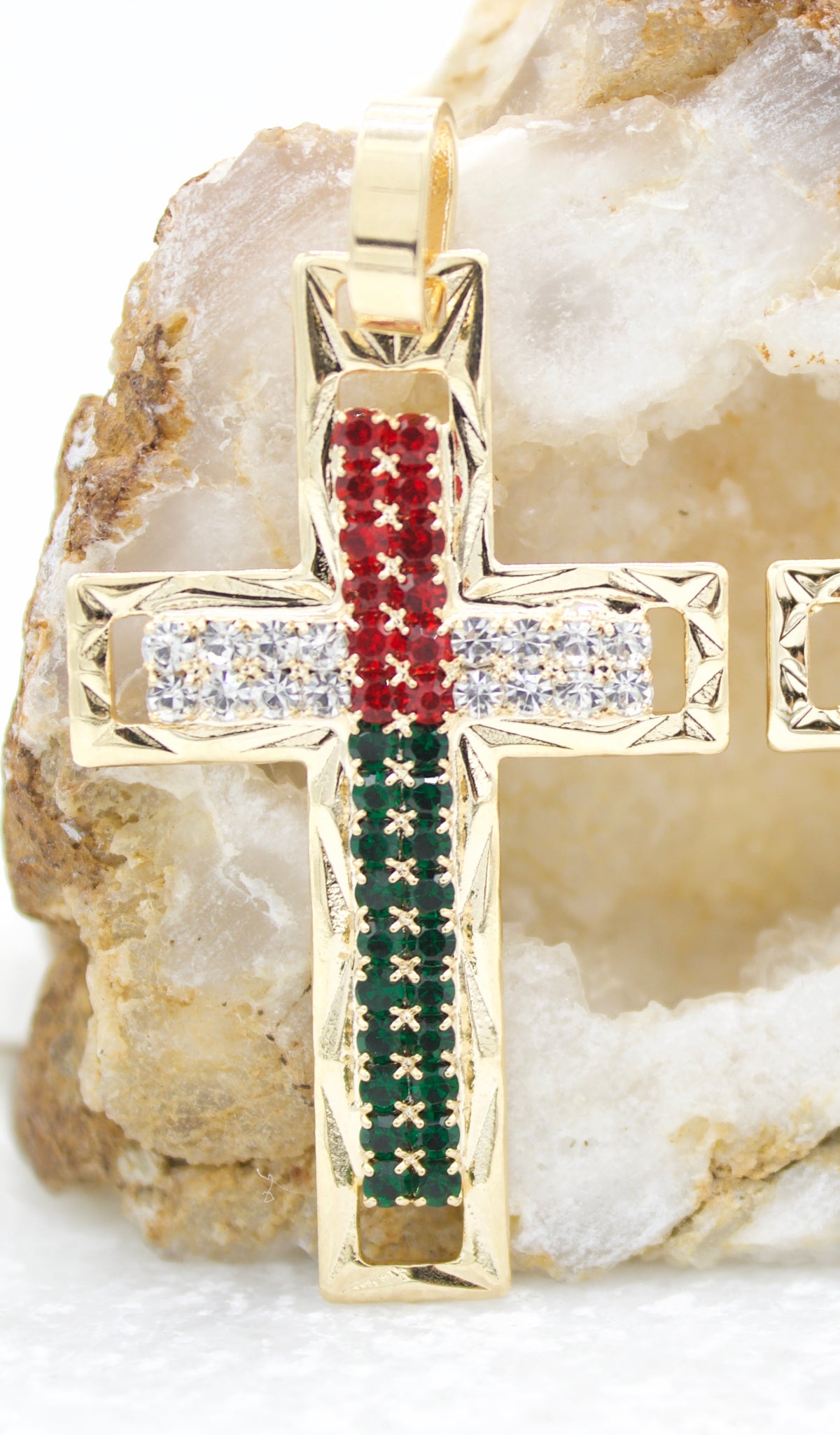 A gold cross pendant encrusted with red, white, and green gemstones rests against a natural crystal geode.