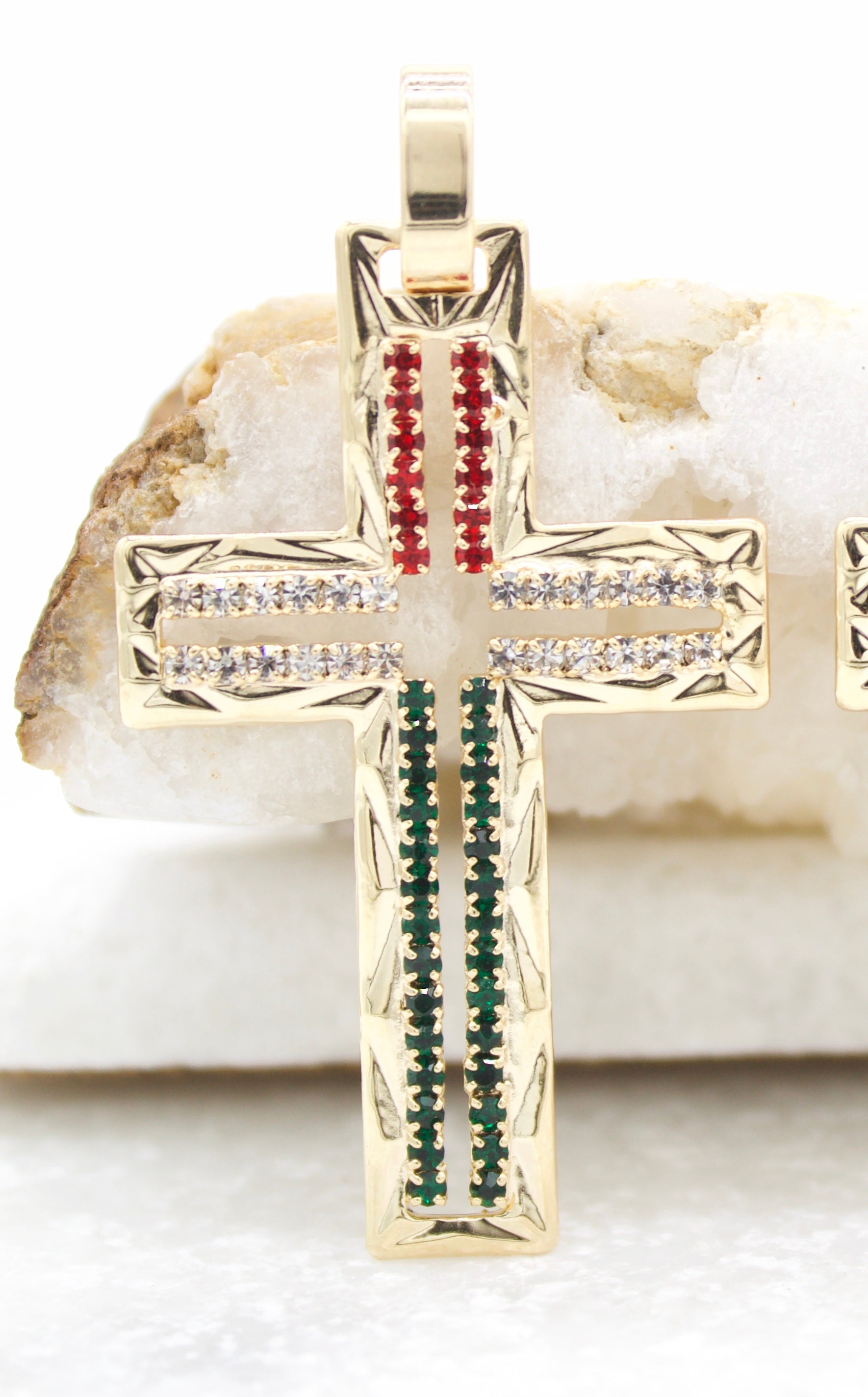 A gold cross pendant encrusted with red, white, and green gemstones rests against a white stone.