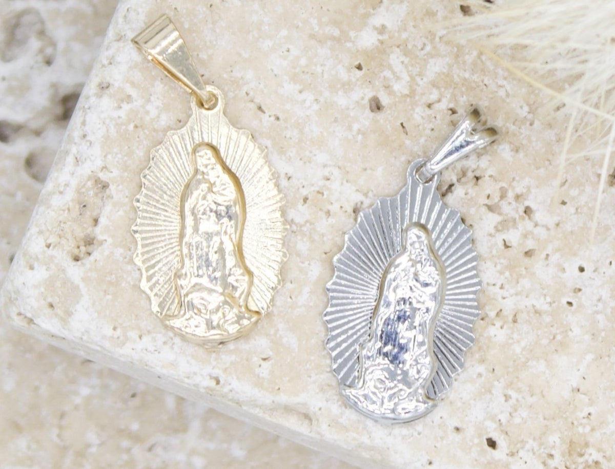 Two Our Lady of Guadalupe pendants, one in gold and one in silver, rest side by side on a textured stone surface.