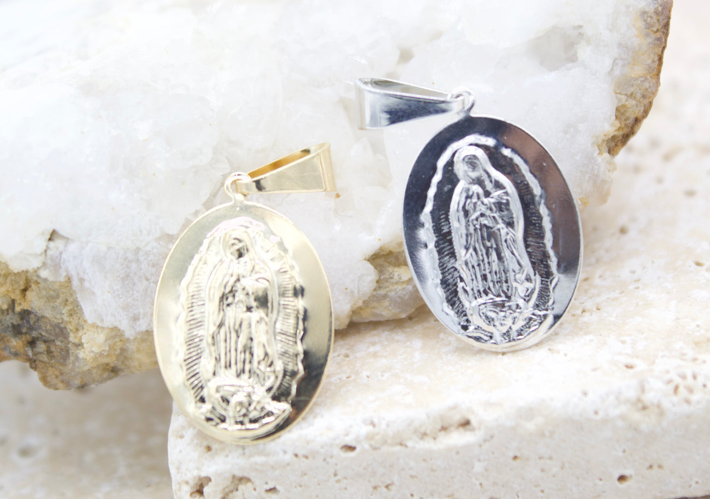 A gold and a silver Virgin Mary pendant rest on a white crystal and stone surface.