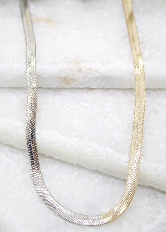 A sleek two-tone herringbone chain necklace rests on a white marble surface, blending silver and gold links.
