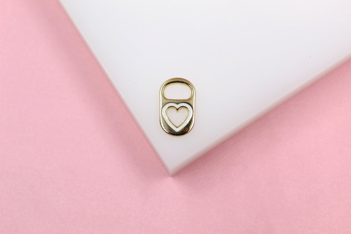 A gold soda tab charm with white enamel heart inlay rests on a white display block against a soft pink background.