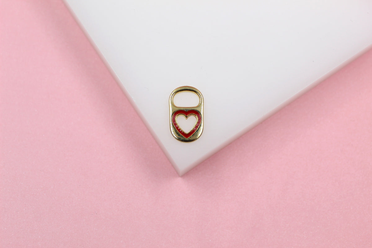A gold soda tab pendant featuring a red enamel heart rests on a white platform against a pink background.