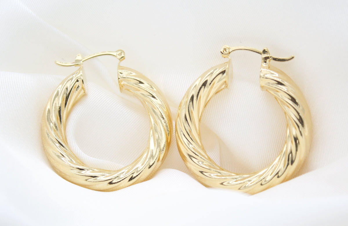 A pair of polished gold twist hoop earrings rests gracefully on a white silk background.