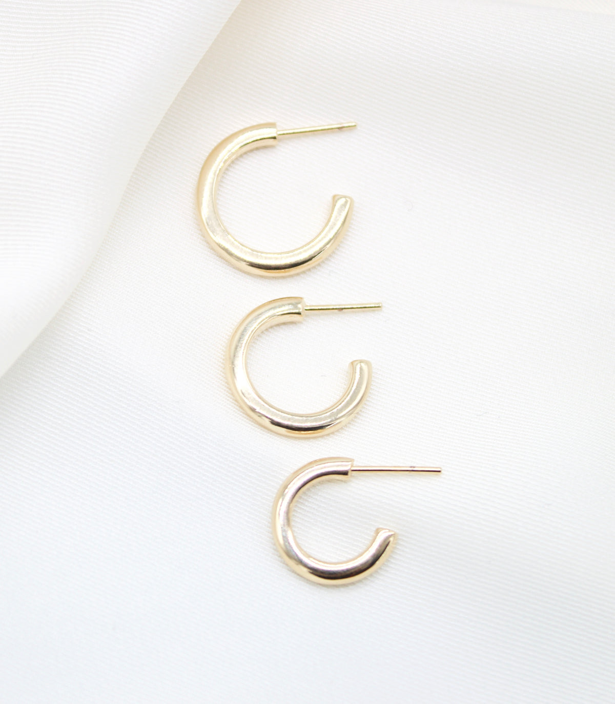 Three graduated gold hoop earrings rest neatly on a soft white silk background.