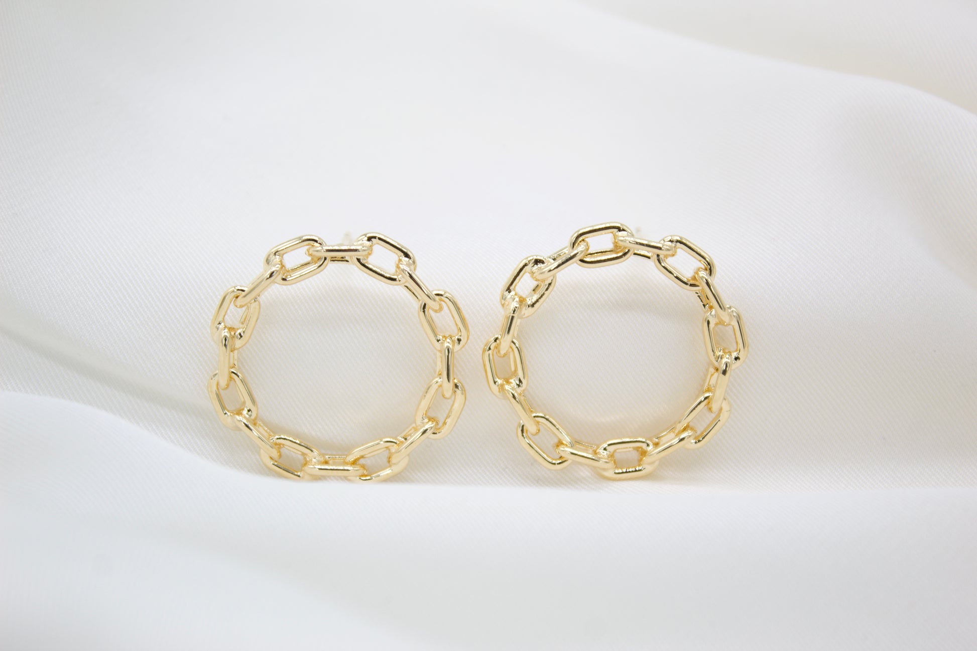 Two round gold chain-link hoop earrings rest on a white silk background.
