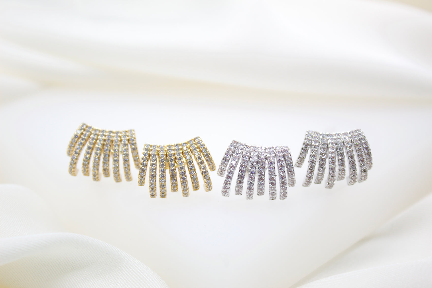 Four multi-strand pavé diamond earrings in gold and silver sparkle against a soft white silk background.