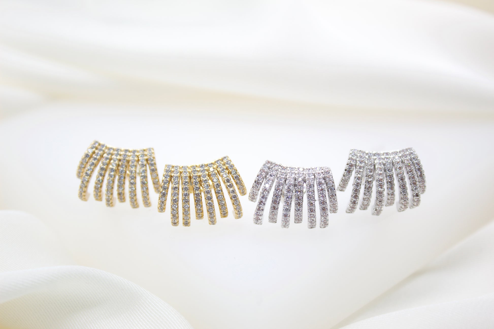Four multi-strand pavé diamond earrings in gold and silver sparkle against a soft white silk background.