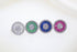 Four round sterling silver cluster rings sparkle on a white surface, each featuring a central halo of pave-set diamonds surrounded by baguette stones in clear, green, blue, and pink.