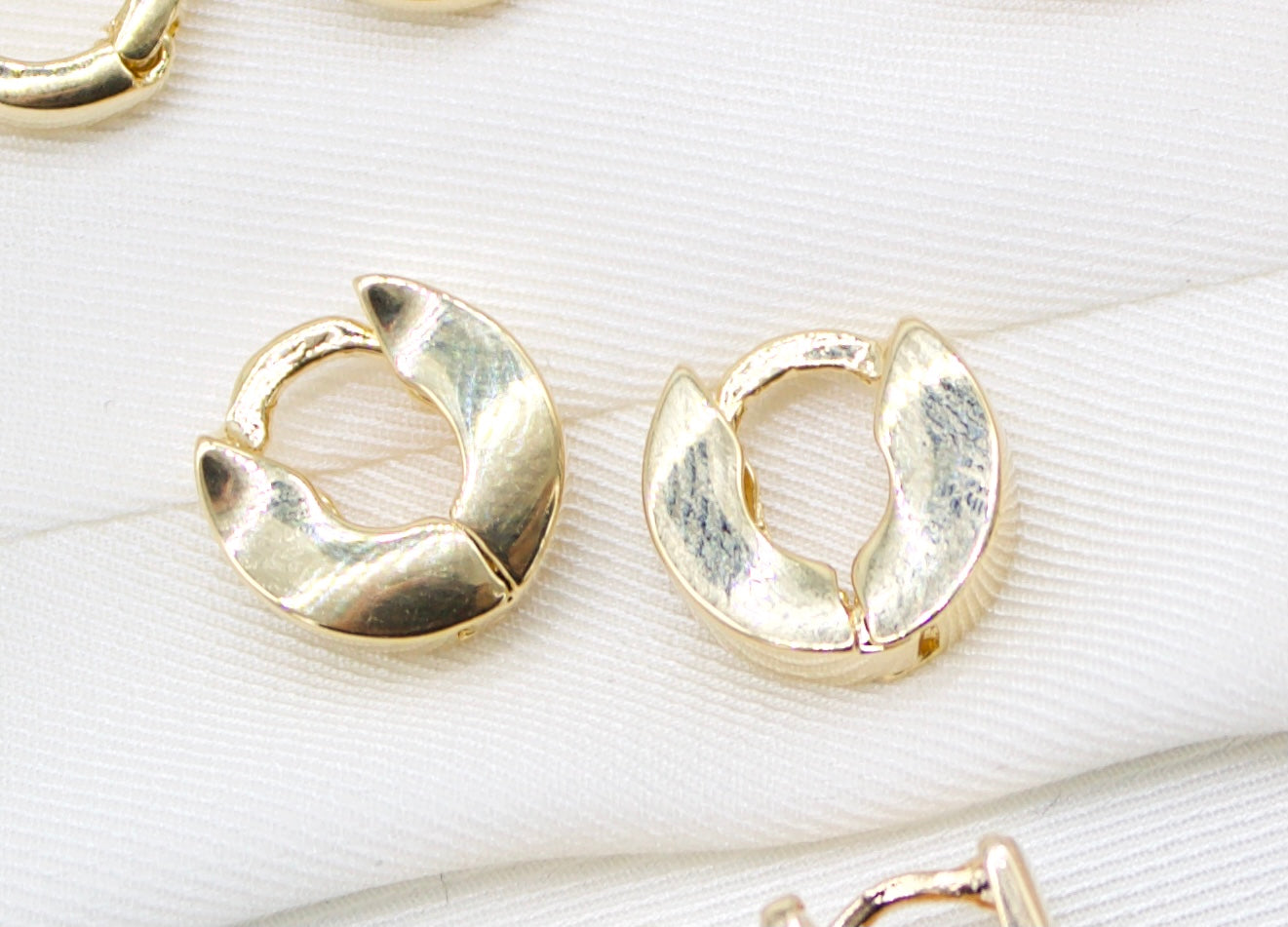 Two small gold huggie hoop earrings with a textured design rest on a white fabric surface.