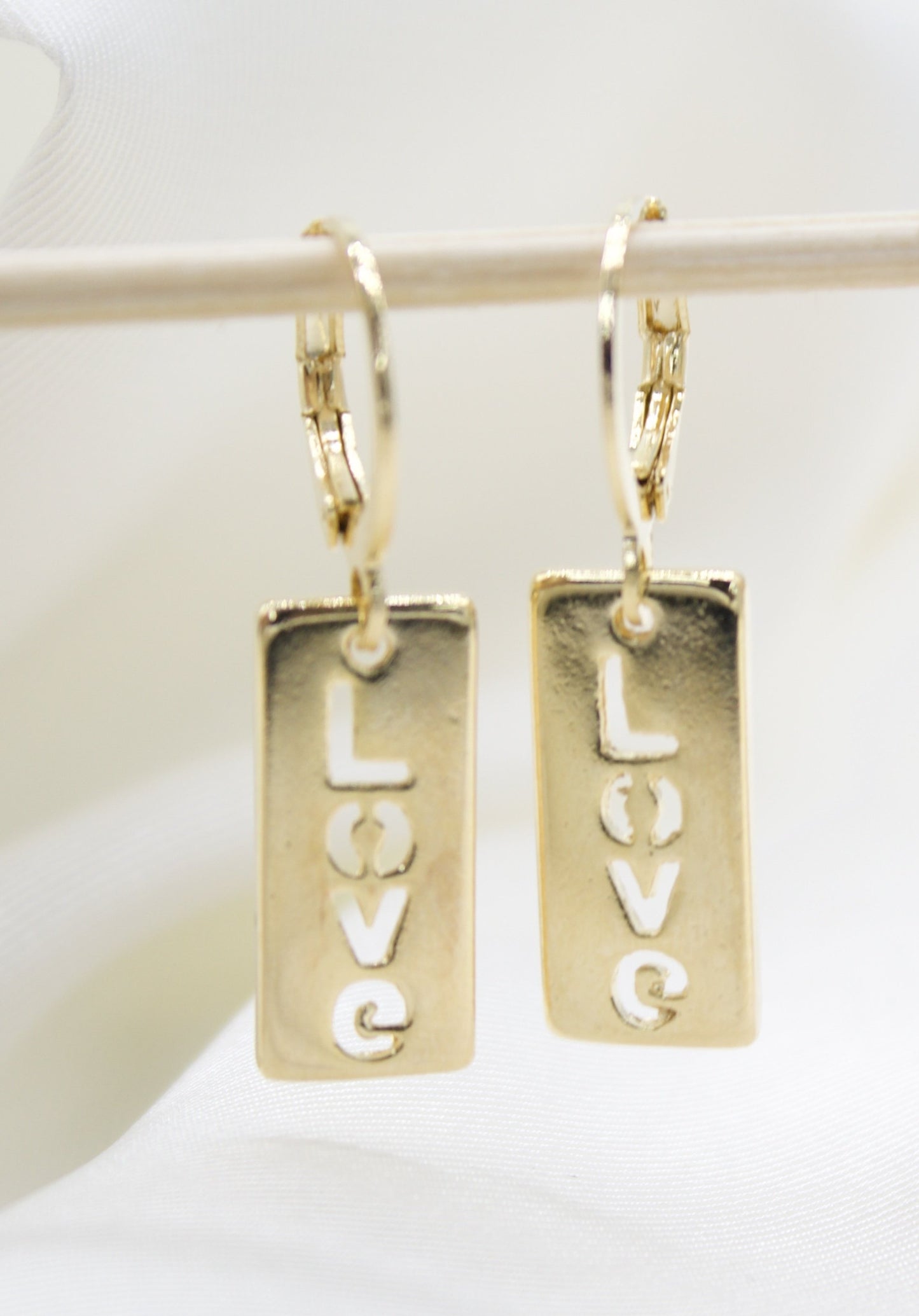 A pair of gold rectangular dangle earrings featuring the word "LOVE" engraved vertically hang delicately from a wooden stick against a soft, white background.