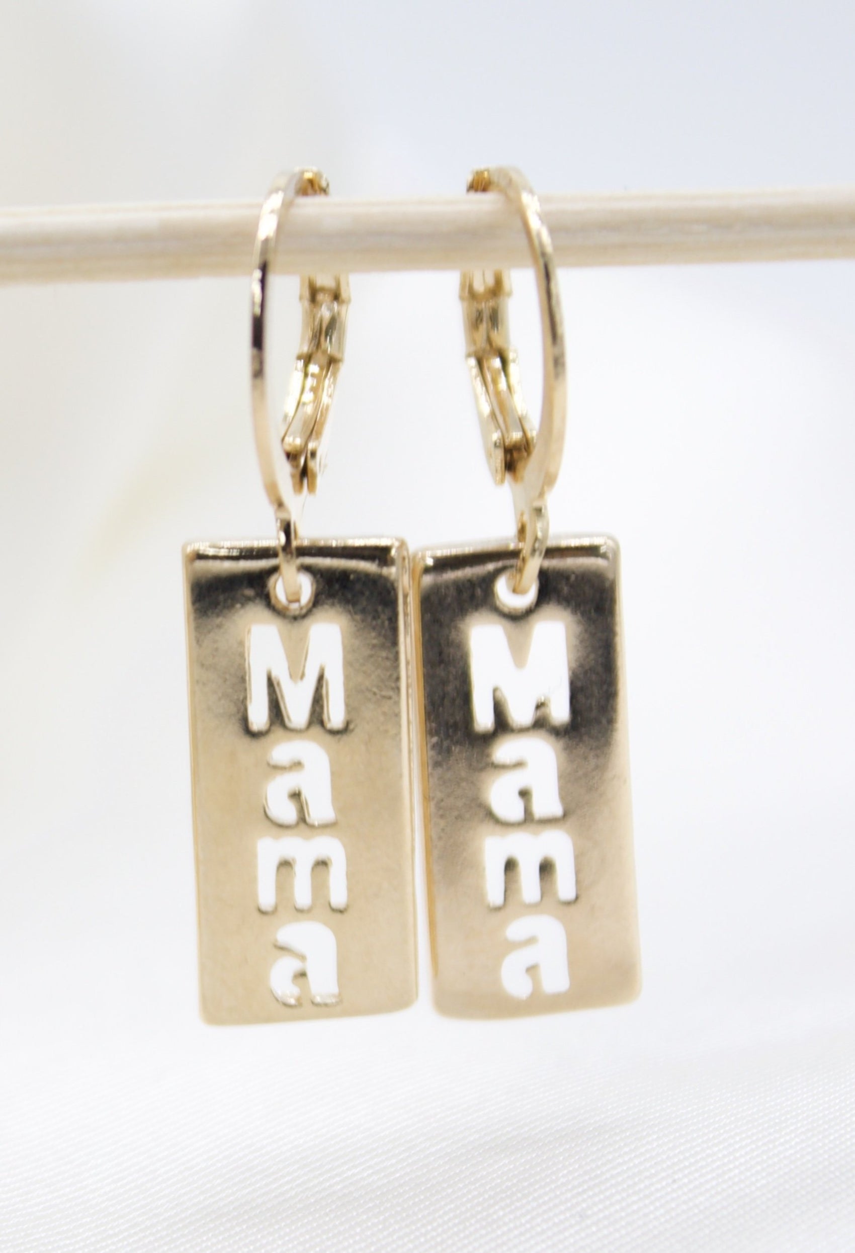 A pair of gold hoop earrings with rectangular pendants featuring the cut-out word "Mama" hangs against a soft white background.