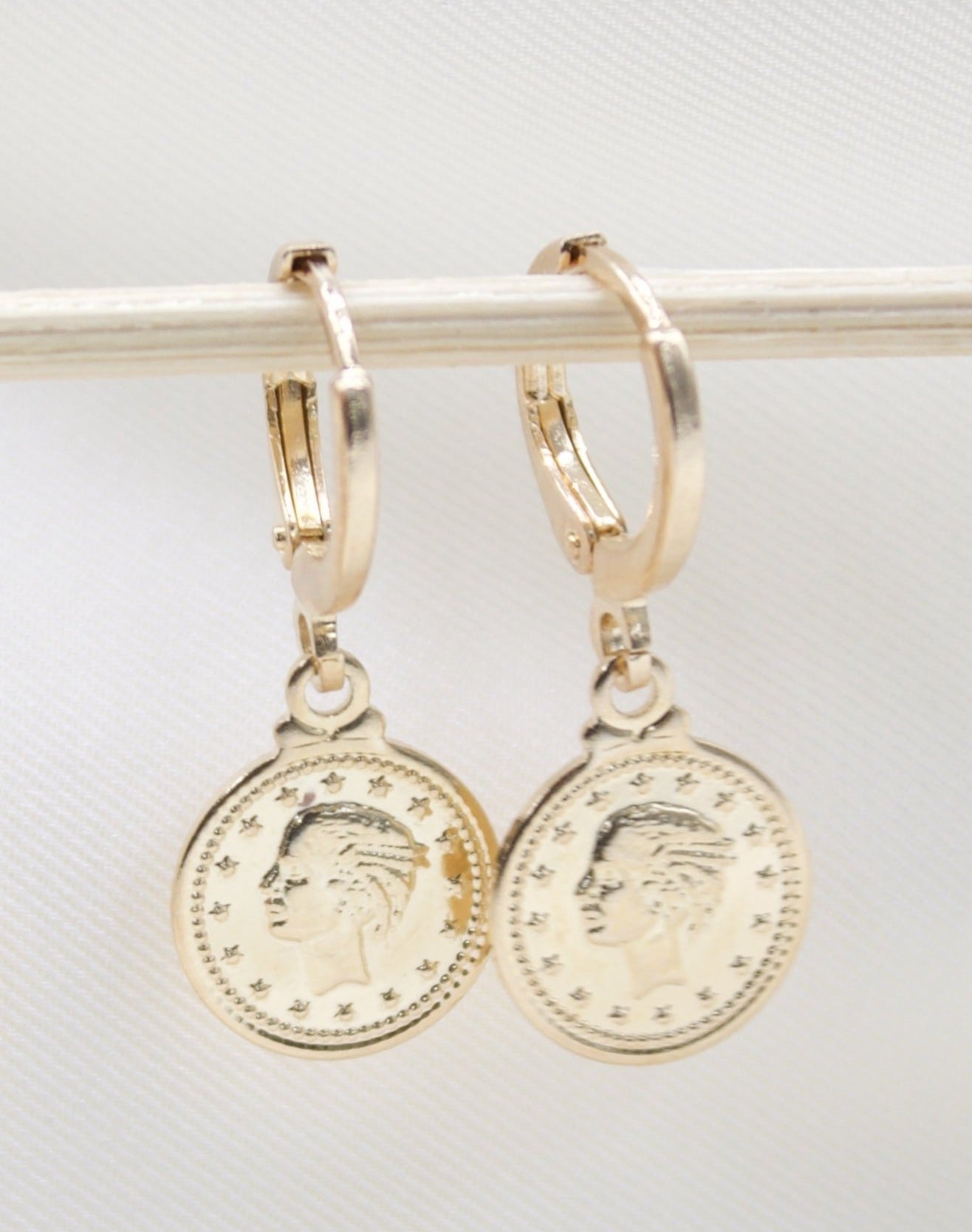 A pair of gold coin hoop earrings hang from a white rod against a neutral background.