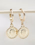 A pair of gold coin hoop earrings hang from a white rod against a neutral background.