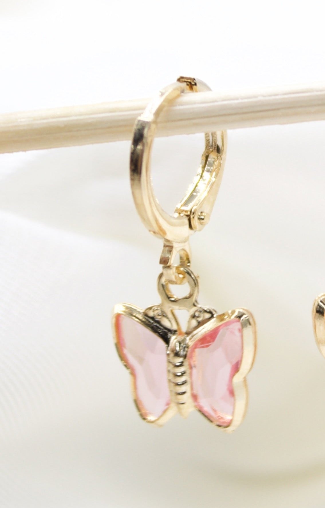 A delicate gold hoop earring hangs from a wooden stick, featuring a charming pink crystal butterfly pendant.