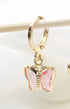 A delicate gold hoop earring hangs from a wooden stick, featuring a charming pink crystal butterfly pendant.