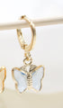 A gold hoop earring hangs from a thin wooden stick, featuring a delicate blue butterfly charm dangling below.