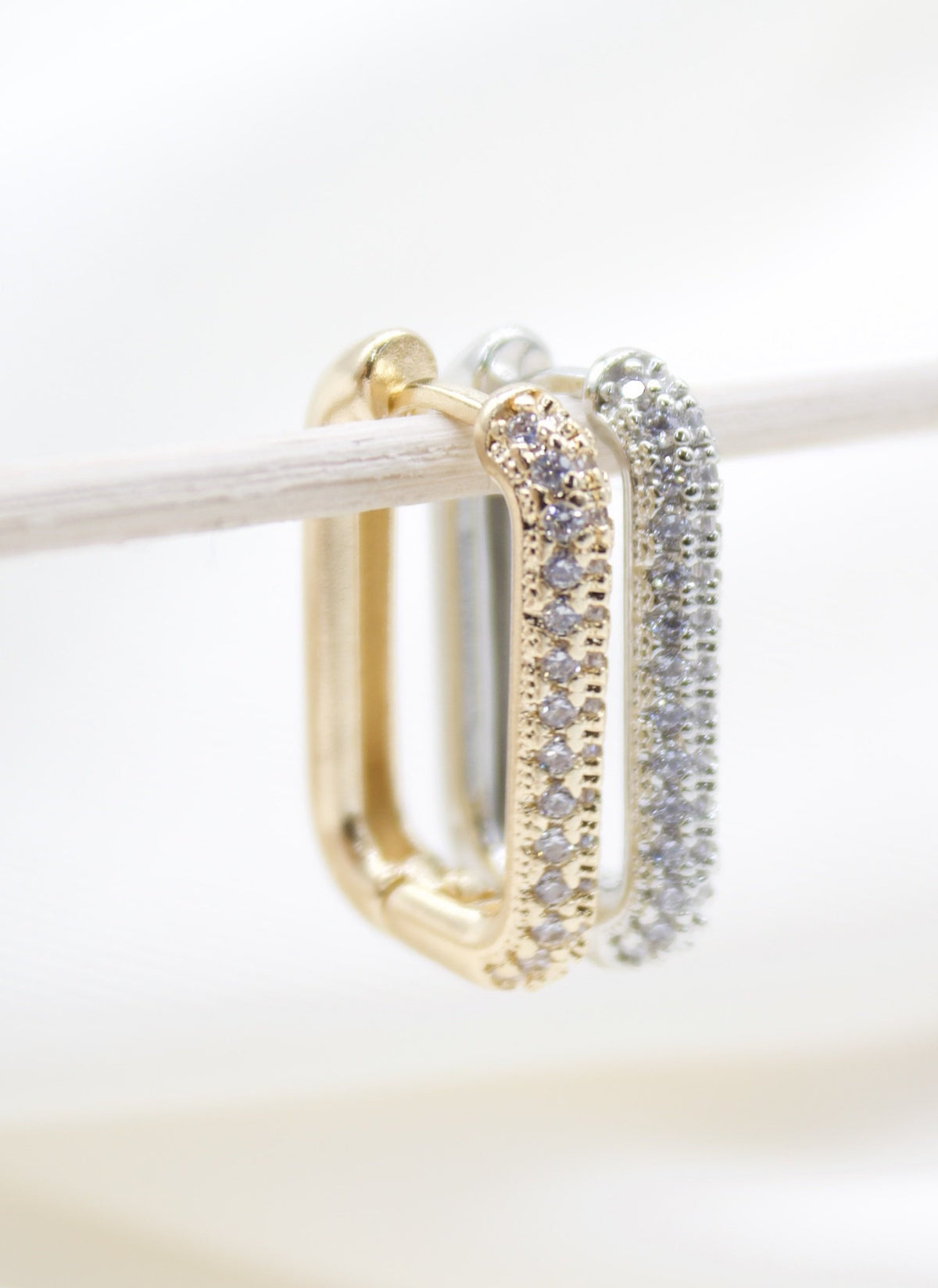 Two rectangular pave huggie hoop earrings, one gold and one silver, hang delicately from a wooden stick against a bright, neutral background.
