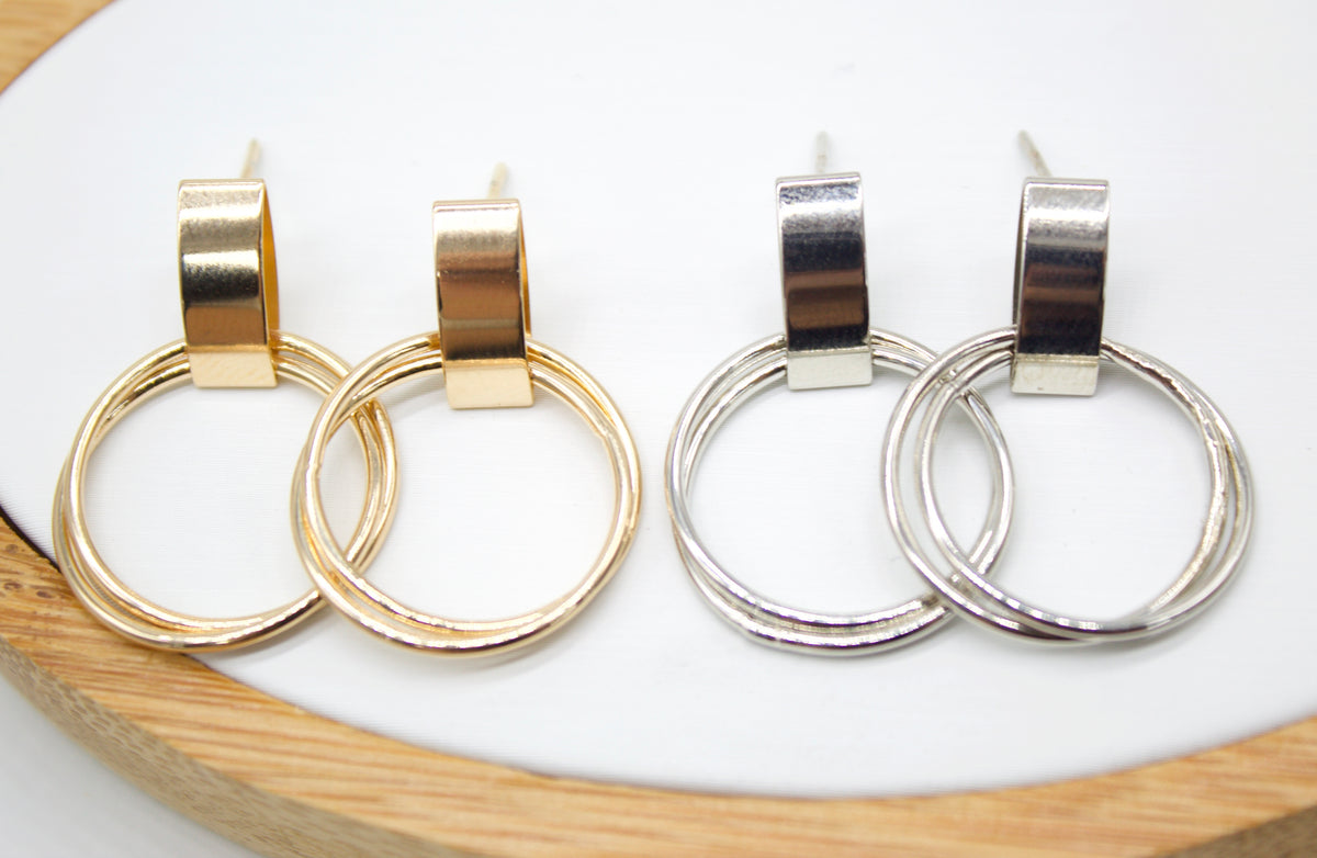 Two pairs of geometric double hoop earrings, one gold and one silver, rest side by side on a white surface.