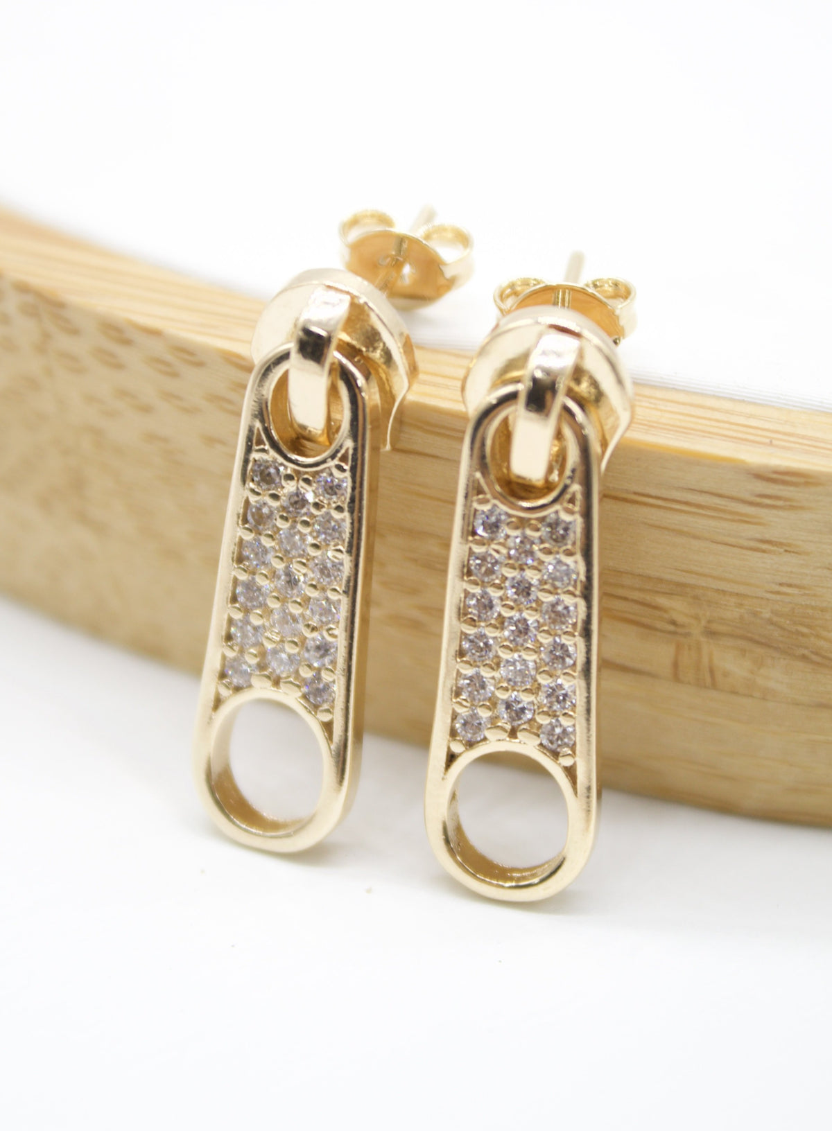 A pair of gold zipper pull earrings encrusted with sparkling cubic zirconia stones rests against a wooden display.