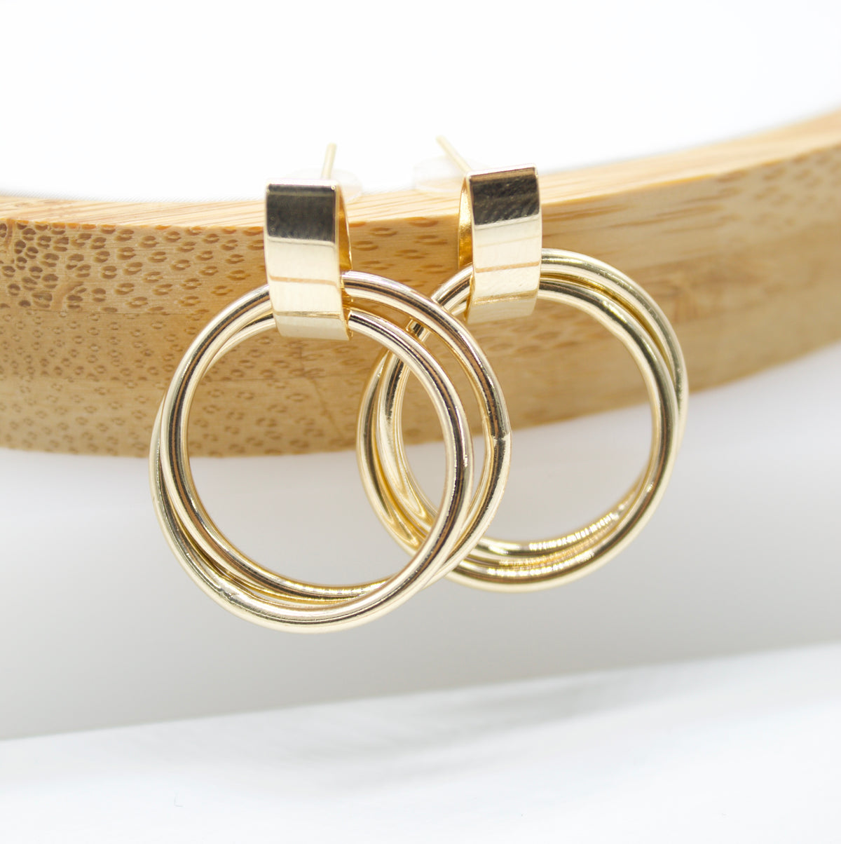 A pair of polished gold double-hoop earrings rests against a curved wooden display.