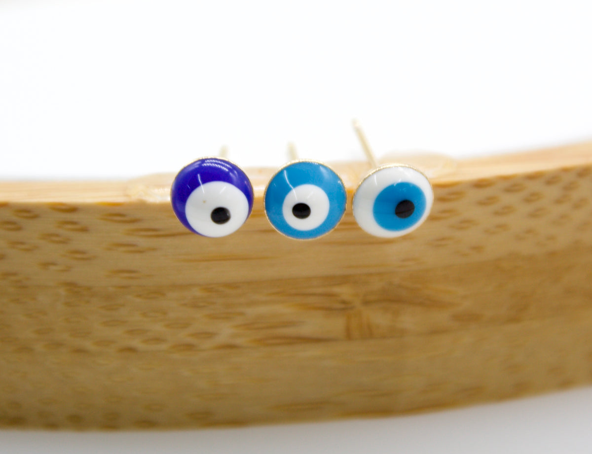 Three matching evil eye stud earrings in dark blue, light blue, and white rest on a curved wooden surface.