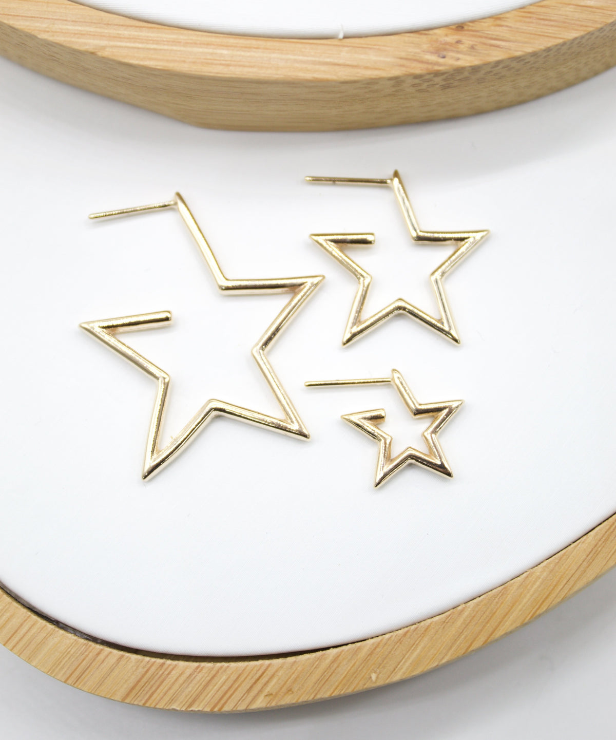 Three gold star-shaped hoop earrings of varying sizes rest on a white display surface inside a wooden embroidery hoop.