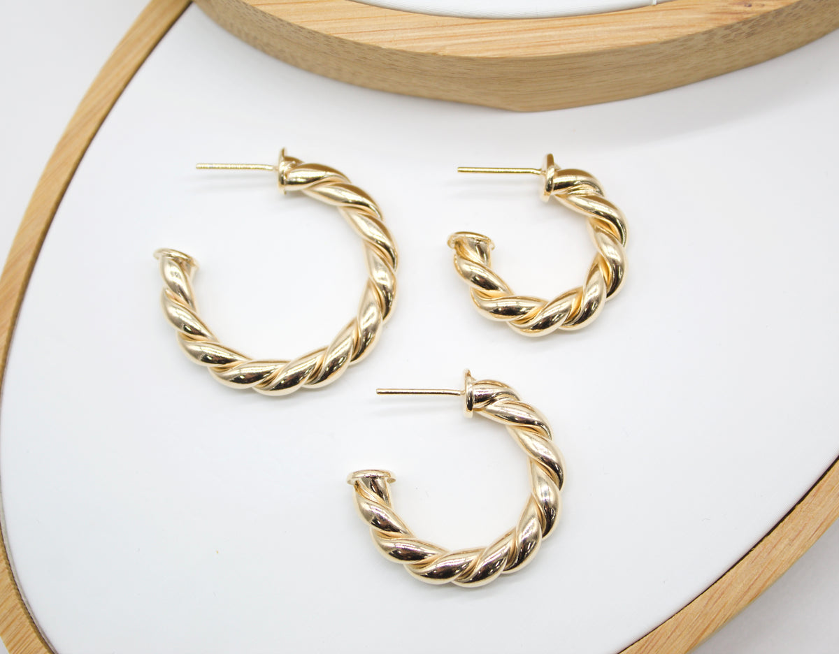 Three polished gold twisted hoop earrings rest on a white surface inside a wooden display tray.