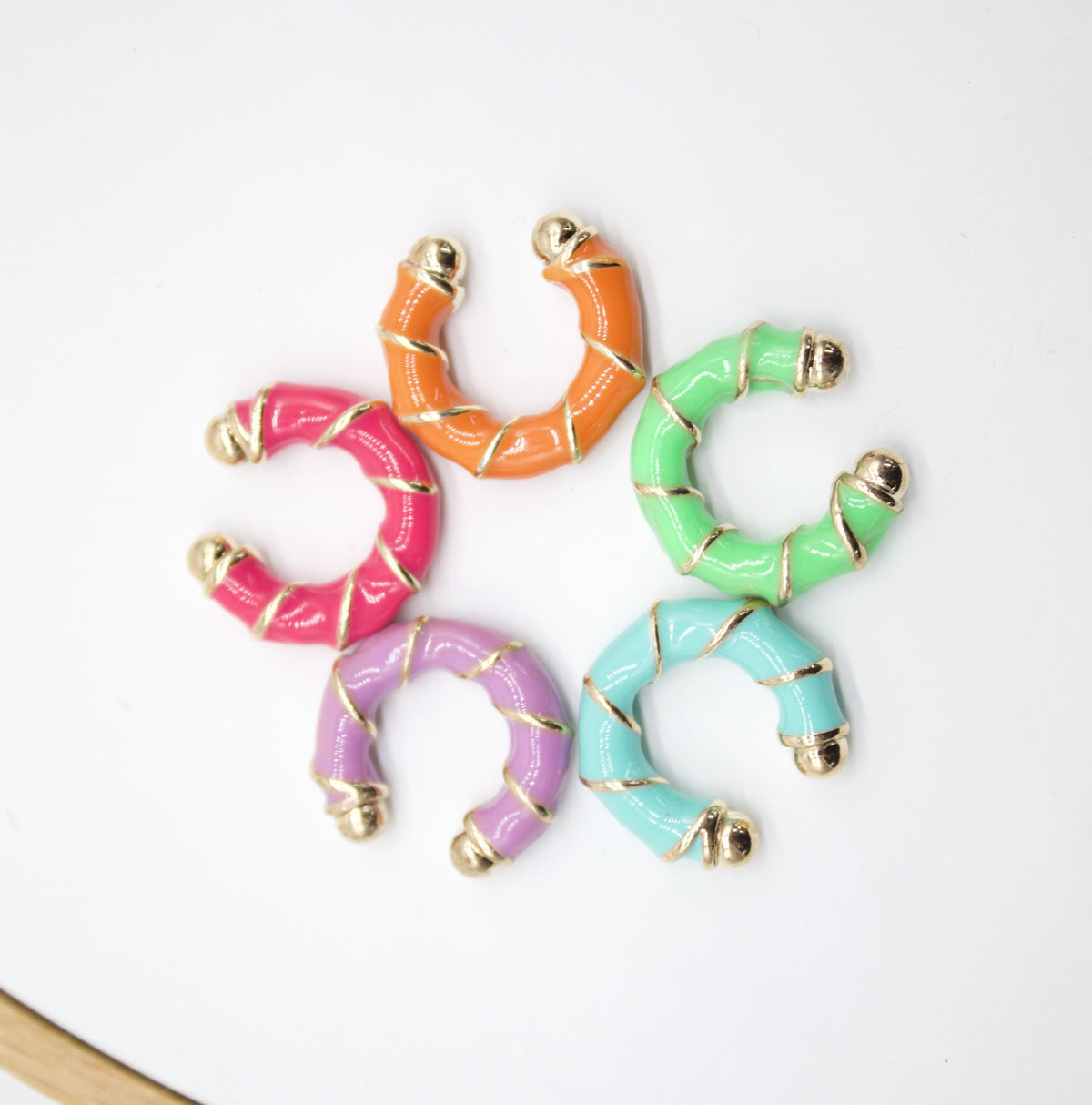 Five colorful enamel and gold-plated huggie earrings are arranged in a circle on a white background.