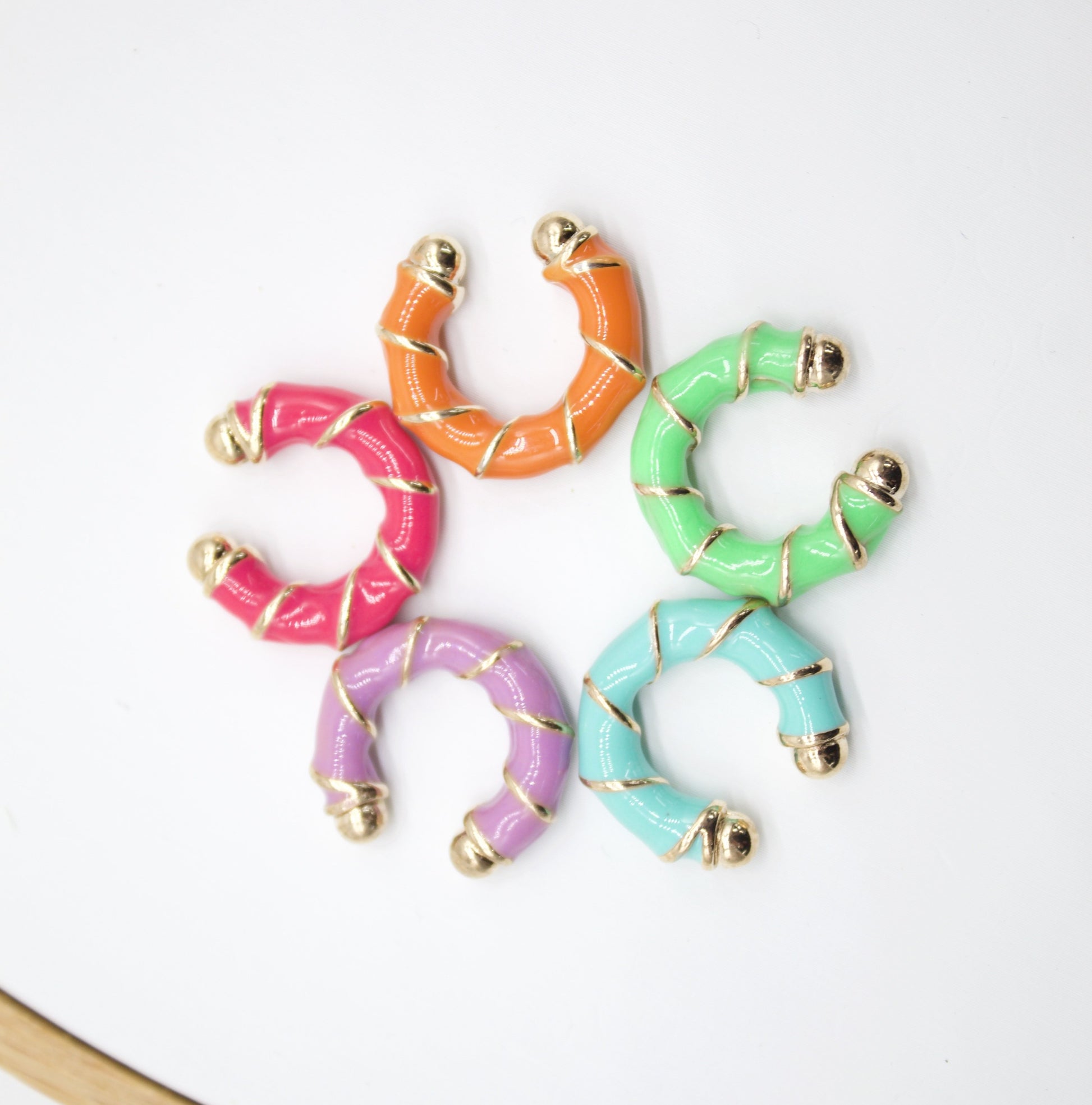 Five colorful enamel and gold-plated huggie earrings are arranged in a circle on a white background.