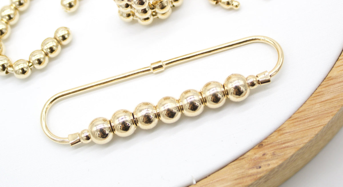 A gold safety pin brooch embellished with a row of shiny metallic beads rests on a white surface next to matching jewelry pieces.