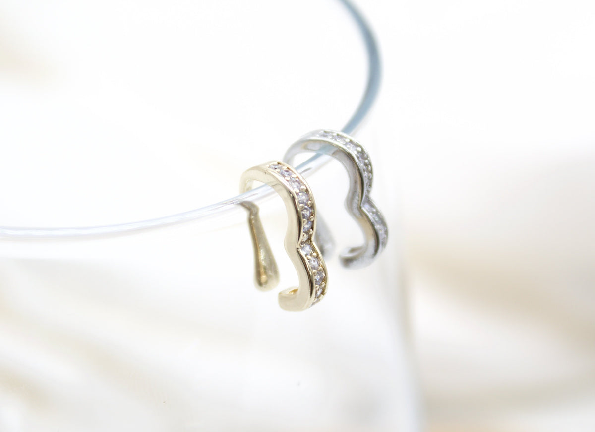 Two shimmering heart-shaped ear cuffs, one in gold and one in silver pavé-set with crystals, rest delicately on a transparent display loop against a soft white background.