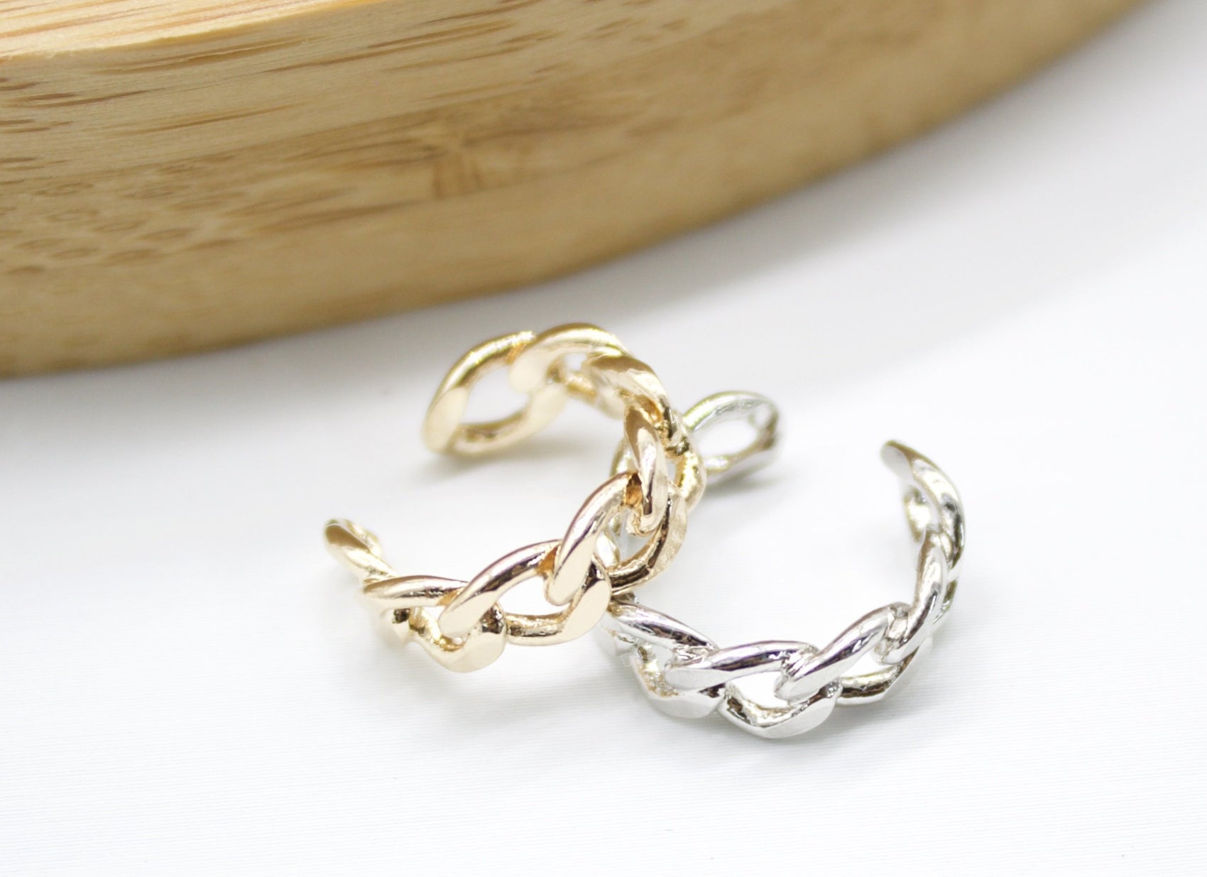 Two interlocking chain-link ear cuffs, one gold and one silver, rest on a clean white surface beneath a wooden display.