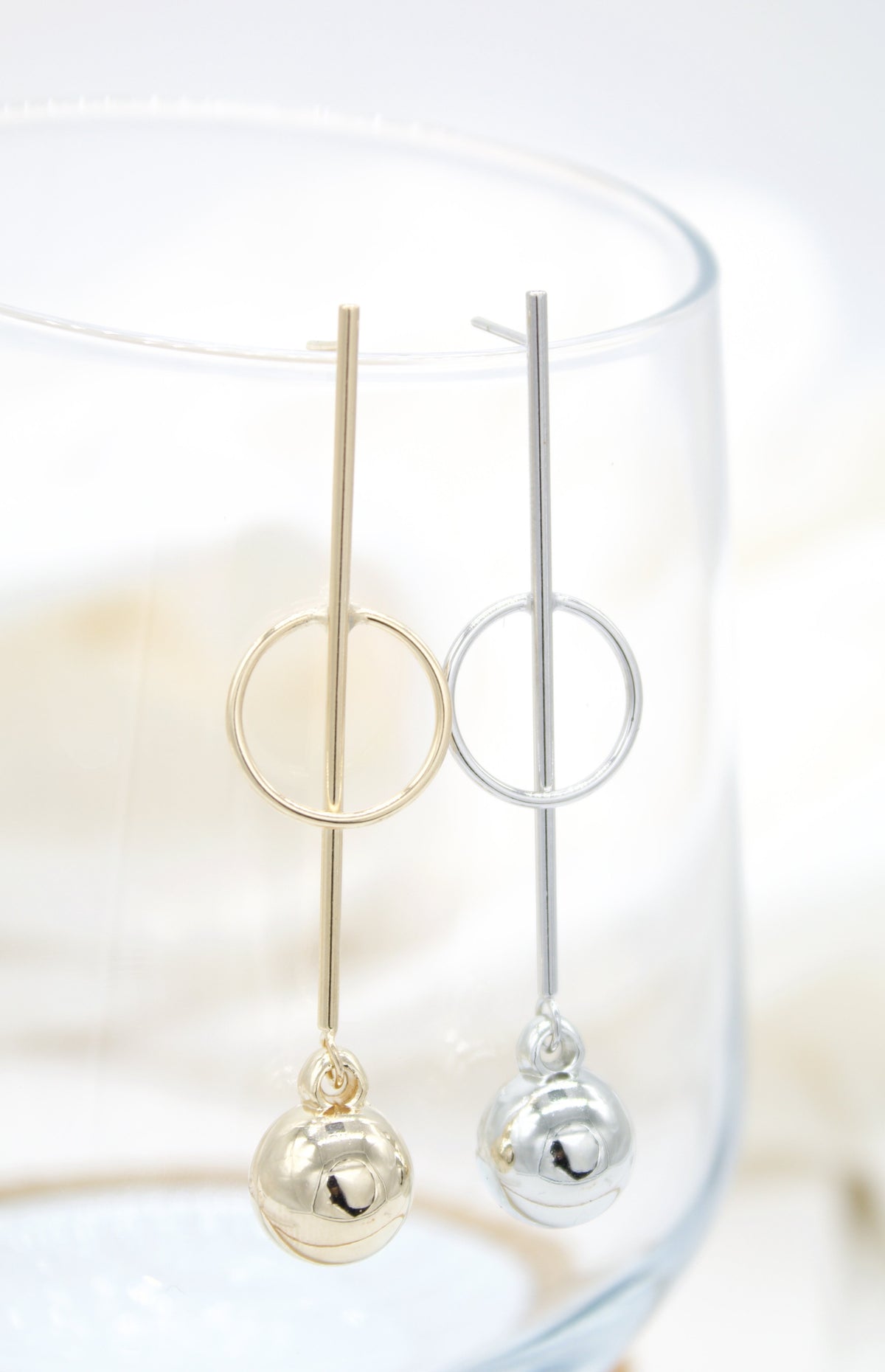 A gold and a silver geometric drop earring featuring a vertical bar, a circle, and a dangling ball hang delicately on the rim of a clear glass.